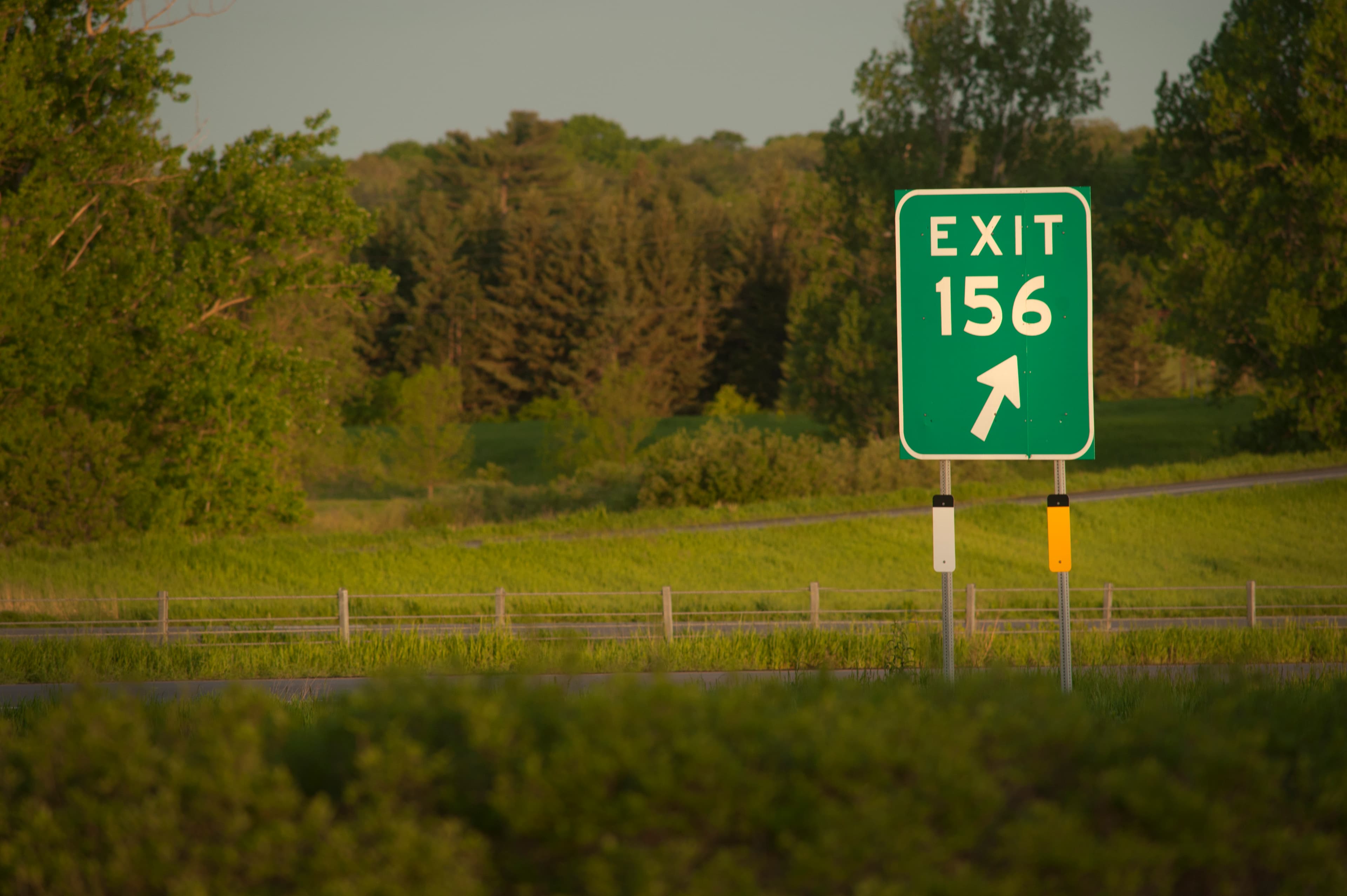 Highway Exit 156 sign at golden hour beside a green field and treeline